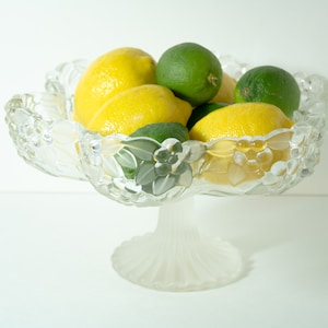 May include: A clear glass pedestal bowl with a floral design, filled with lemons and limes.