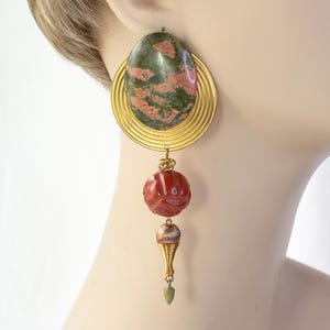 May include: A single earring featuring a large, oval-shaped stone with green and pink hues, set in a gold-toned, spiral-edged frame. Below, a carved red bead and a gold-toned cone-shaped element dangle, creating a unique, eye-catching design.