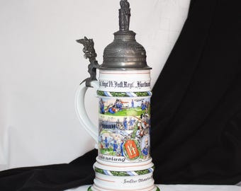 May include: A white ceramic beer stein with a pewter lid and a pewter figure of a soldier on top. The stein is decorated with a scene of soldiers in a field and the text "1. Inf. Regt. Harl. Erinnerung" in German. The stein is approximately 12 inches tall.