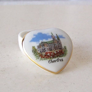 May include: A white porcelain heart-shaped trinket box with a gold trim. The box features a painted image of the Chartres Cathedral in France with a blue sky and red flowers in the foreground. The text "Chartres" is printed in black below the image.