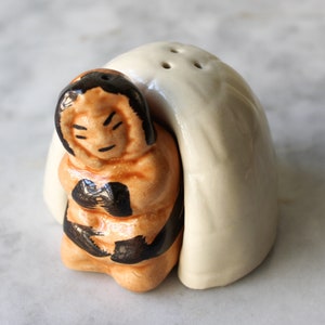 Op de afbeelding: Keramische zout- en peperstrooierset met een bruin en zwart beeldje van een persoon die een gekapte kleding draagt, die een iglo voorstelt. De iglo is wit met drie gaten bovenop.