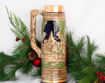 May include: A ceramic beer stein with a detailed relief of a castle scene, labelled "SCHLOSS ANHOLT". The stein has a wooden-look handle and lid, with green, brown, and gold accents. The background includes evergreen branches and red berries.