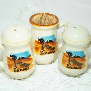 May include: Set of three white ceramic salt and pepper shakers with a toothpick holder. Each shaker has a brown, orange, and blue scene of a windmill and farm with the word "Kansas" printed on the front.