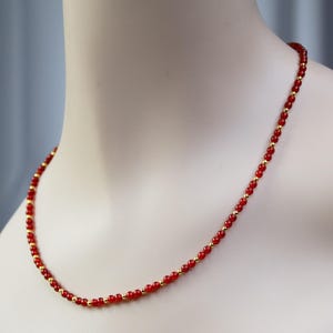 May include: A necklace featuring a repeating pattern of small, round, vibrant red beads interspersed with tiny, polished gold-tone metallic spheres. The beads are uniform in size and arranged in a single strand that rests against the neck.