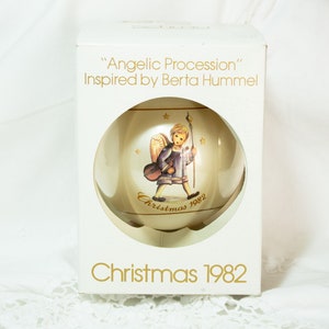 Può includere: Un addobbo natalizio bianco con un bordo dorato e un'immagine di un angelo che suona uno strumento musicale. L'addobbo è in una scatola bianca con il testo "Angelic Procession" Inspired by Berta Hummel e "Christmas 1982".