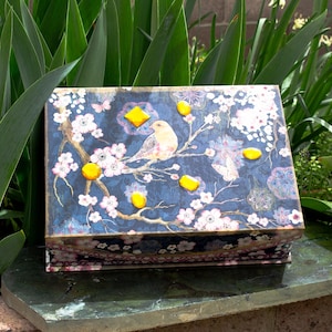 May include: A decorative wooden box with a blue background and a floral design. The box has a hinged lid and is decorated with yellow accents and a small bird.