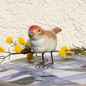 Puede incluir: Una figurita de pájaro de cerámica con la cabeza roja, el cuerpo marrón y blanco y el pico amarillo. El pájaro está posado en una mesa con una tapa de mosaico.