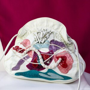 May include: A white leather pouch bag featuring a gathered top closure and a decorative white leather tassel. The exterior is adorned with colorful, textured patches in shades of pink, red, purple, and teal, accented by a white braided leather strap.