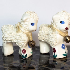 May include: Two white ceramic lambs with blue eyes, red lips, and gold bells. The lambs have a fluffy white texture and are decorated with blue, red, and green flowers.