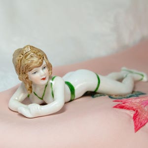 Könnte beinhalten: Eine Porzellanfigur einer Frau, die auf dem Bauch liegt und einen weißen Badeanzug mit grünem Besatz trägt. Die Figur steht auf einer rosa Oberfläche.