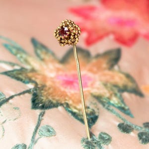 May include: A gold pin with a pink gem in the centre, surrounded by gold beads. The pin is on a pink and green floral fabric.