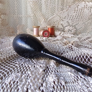 Może przedstawiać: Czarny drewniany młotek z gładką, zaokrągloną główką i długim, smukłym trzonkiem. Młotek leży na białym, szydełkowym obrusie. W tle widać trzy szpulki nici, jedną różową, jedną czerwoną i jedną białą.