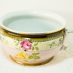 Pode incluir: Um penico de porcelana branca com um desenho floral rosa e amarelo. O penico tem uma borda dourada e uma alça.