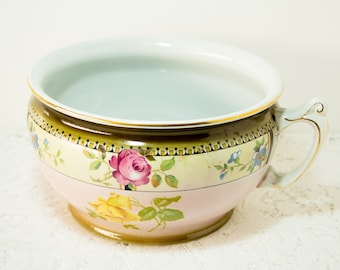 May include: A white porcelain chamber pot with a pink and yellow floral design. The pot has a gold rim and a handle.