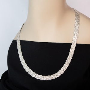 May include: A silver-colored, braided necklace. The necklace is draped around the neck and chest, resting on a black top. The braided design gives the necklace a textured appearance. The necklace is a statement piece, suitable for various occasions.