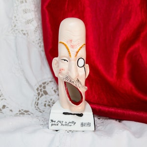 May include: A white ceramic figurine of a man with a mustache and a monocle. He is wearing a bow tie and has a large open mouth. The figurine is on a white base with the text "For He's a jolly good fellow..." written on it.