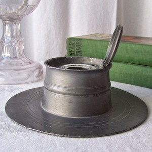 May include: A pewter inkwell with a hinged lid, the lid is open. The inkwell has a round base and a cylindrical body. The inkwell is sitting on a white surface.