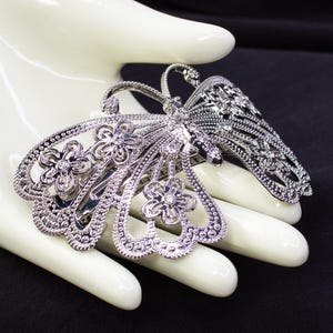 May include: A silver-toned butterfly hair clip with intricate detailing. The clip features openwork wings and is adorned with small, sparkling embellishments. The butterfly's body and wings are decorated with floral patterns and tiny crystals, creating a delicate and elegant accessory.