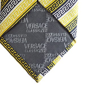 Puede incluir: Una corbata de seda negra con un patrón de llave griega amarillo y blanco. La corbata está forrada con una tela negra que tiene las palabras "VERSACE CLASSIC" repetidas en blanco.