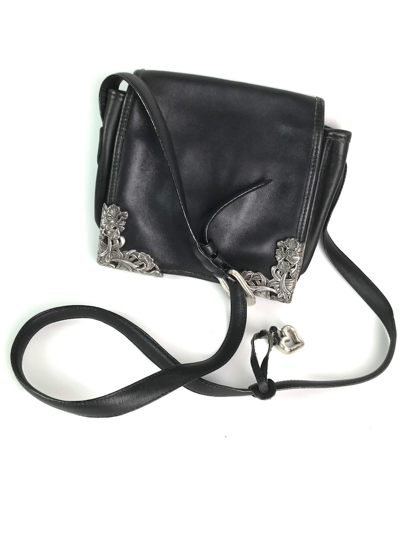 Może przedstawiać: Czarna sk&oacute;rzana torba na ramię ze srebrnymi, kwiatowymi narożnikami i zawieszką w kształcie serca. Torba posiada klapę i regulowany pasek. Torba ma szerokość około 20 cm.