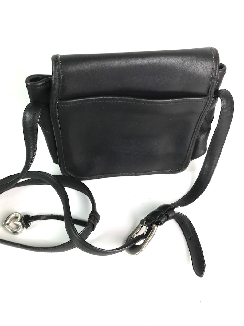 Może przedstawiać: Czarna sk&oacute;rzana torba na ramię z klapką i przednią kieszenią. Torba posiada długi, regulowany pasek ze srebrną klamrą i zawieszką w kształcie serca.