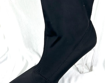 May include: A black, over-the-knee boot with a pointed toe and a clear, block heel. The boot is made of a stretchy material and has a sleek, modern design. The heel is approximately 3 inches (7.6 cm) high.