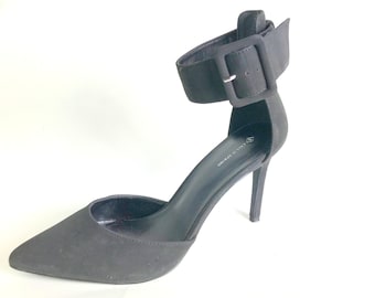 May include: Black suede heeled pumps with an ankle strap and buckle closure. The shoes have a pointed toe and a stiletto heel.