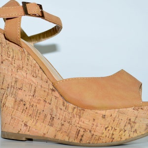 Puede incluir: Sandalias de cuña de color marrón claro con plataforma de corcho y correa de tobillo con hebilla dorada.