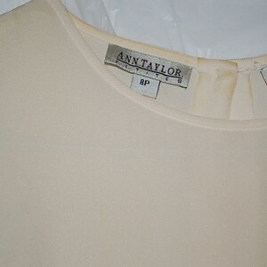 May include: Cream-colored Ann Taylor Petite top. The top features a round neckline and short sleeves. The Ann Taylor label is visible at the back of the neck, with the size 8P. The fabric appears to be a lightweight material.