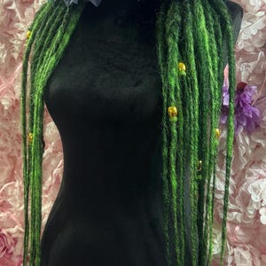 Könnte beinhalten: Ein schwarzer Mannequinkopf zeigt grüne Dreadlocks mit goldenen Perlen und dunkelblauen Blumendekorationen. Die Dreadlocks fallen herab und bilden einen Kontrast zu der schwarzen Form und dem rosa Blumenvorhang. Die Dreadlocks sind leuchtend grün.