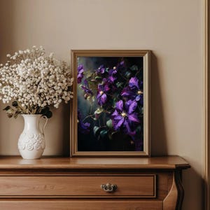May include: A framed floral print featuring vibrant purple clematis flowers with yellow centers and green leaves. The artwork is set against a dark background, framed in gold, and displayed on a wooden dresser. A white vase with white flowers is on the left.