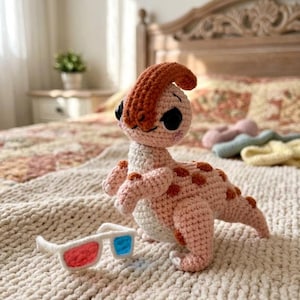 May include: A crocheted dinosaur toy in shades of pink and orange, with large black eyes and a distinctive crest. Beside it are white 3D glasses with red and blue lenses. The toy is on a textured, off-white surface.