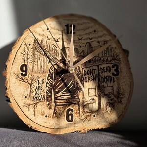 May include: A round wooden clock with a natural bark edge. The clock face features a pyrography design of a figure, trees, and the text "WE ARE ALL NEGAN" and "DON'T OPEN DEAD INSIDE". The clock hands are gold.