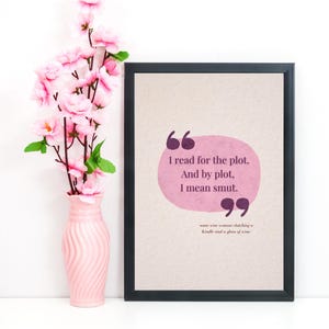 May include: Framed print with the quote "I read for the plot. And by plot, I mean smut." The text is in a pink bubble on a beige background, framed in black. A pink vase with pink flowers is to the left.