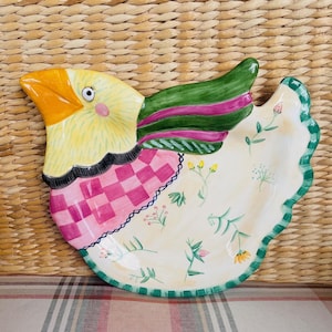 May include: A decorative ceramic chicken-shaped serving platter. The platter features a yellow head with an orange beak, a pink and red checkered body, and green wings with pink stripes. The rim is scalloped and painted green.