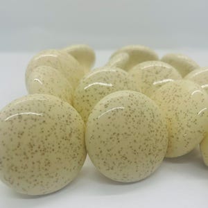 May include: Cream-colored, speckled ceramic knobs. The knobs are round with a glossy finish and a pattern of small, dark brown speckles. The knobs are clustered together, suggesting a set for furniture.