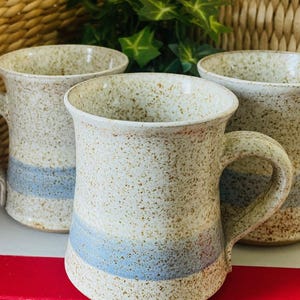 May include: Three speckled ceramic mugs with handles. The mugs have a light beige base color with a blue stripe around the middle. The mugs are sitting on a red book.