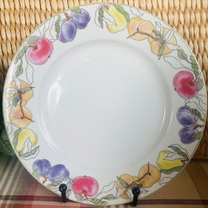 May include: White ceramic plate with a border of colorful fruit illustrations, including apples, plums, and pears. The fruit is painted in shades of red, purple, yellow, and orange, with green leaves. The plate is displayed on a black stand.