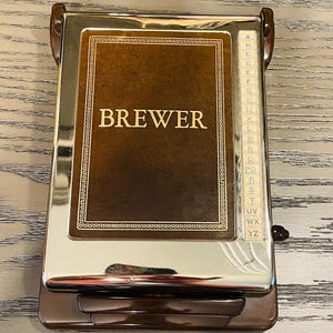May include: A vintage address book with a brown leather cover and the name "BREWER" in gold lettering. The book has a shiny, metallic frame and a lettered index on the side. The book is closed and has a brown plastic base and handle.