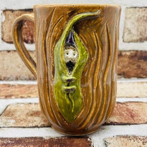 May include: A brown ceramic mug with a textured surface resembling tree bark. A sculpted green pea pod is attached to the front, featuring a small, bearded figure with a dark pointed hat peeking out from the center of the pod.
