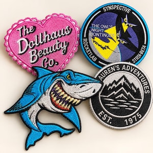 May include: A collection of four embroidered patches: a pink heart-shaped patch with the text The Dollhaus Beauty Co., a circular blue patch with an owl graphic and text Synspective Rocketlab The Owl's Night Continues Strix-Beta, a circular black patch with mountain graphics and text Auren's Adventures Est. 1975, and a blue shark-shaped patch.