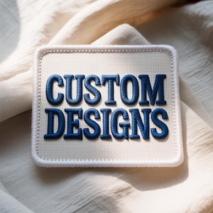 May include: A square patch with the words "CUSTOM DESIGNS" embroidered in blue thread. The patch has a white background and a white border. The patch is on a cream-coloured fabric.