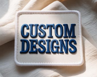 Peut inclure: Un écusson carré avec les mots "CUSTOM DESIGNS" brodés en fil bleu. L'écusson a un fond blanc et une bordure blanche. L'écusson est posé sur un tissu de couleur crème.