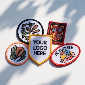 May include: A collection of embroidered patches in various shapes, including round, shield, and oval. The patches feature colorful designs and text, with one displaying "YOUR LOGO HERE". The patches are likely for clothing or accessories.