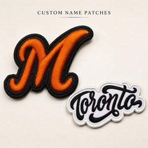 May include: Two embroidered patches with the text CUSTOM NAME PATCHES at the top. One patch is a large orange letter M with a thick black border. The second patch is white with the word Toronto in black cursive script and a white scalloped border.