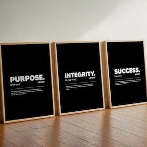 Op de afbeelding: Drie ingelijste zwart-wit prints met de woorden "PURPOSE.", "INTEGRITY." en "SUCCESS." in witte tekst. Elke print heeft een houten lijst en wordt weergegeven op een houten vloer.