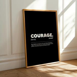May include: A framed black print with the word "COURAGE" in bold white letters. The definition of courage is below the word. The frame is a light brown color, and the print is leaning against a white wall.