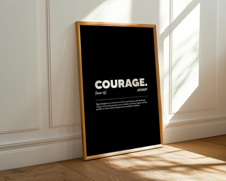 May include: A framed black print with the word "COURAGE" in bold white letters. The definition of courage is below the word. The frame is a light brown color, and the print is leaning against a white wall.