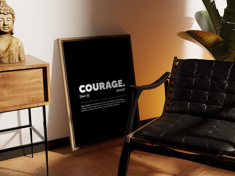 May include: A framed black print with the word "COURAGE" in white, with a definition below. The print leans against a wall next to a wooden side table with a Buddha statue and a black leather chair.