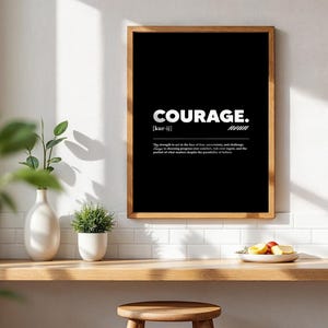 May include: A framed black and white print with the word "COURAGE" in bold white letters. Below the word is the pronunciation and the definition of courage. The frame is a warm brown color, and the print is displayed on a white wall.